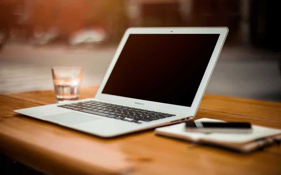 Laptop aperto su un tavolo di legno con un bicchiere d'acqua e un piccolo taccuino