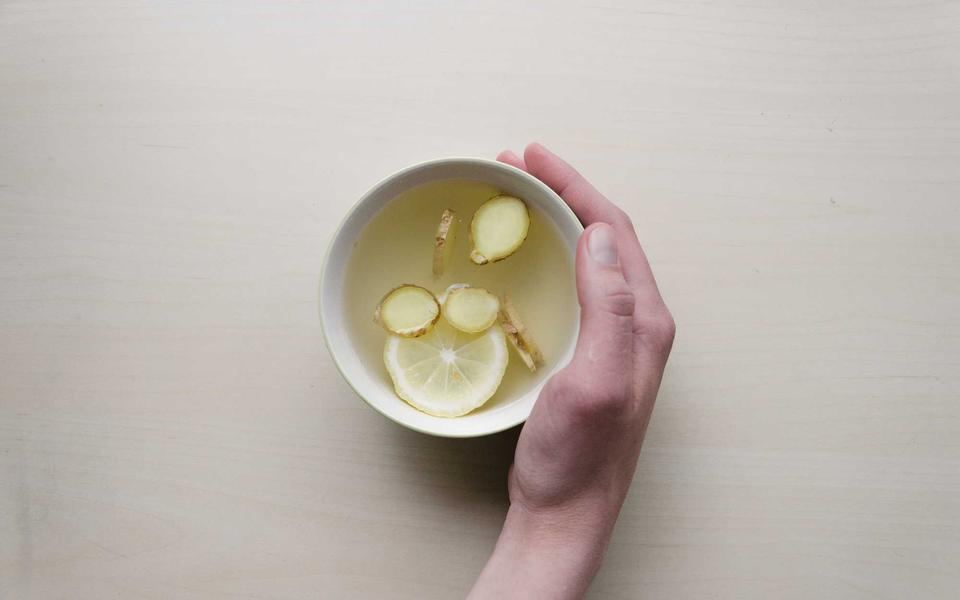 Mano che regge una ciotolina di tisana con una fetta di limone su un tavolo chiaro